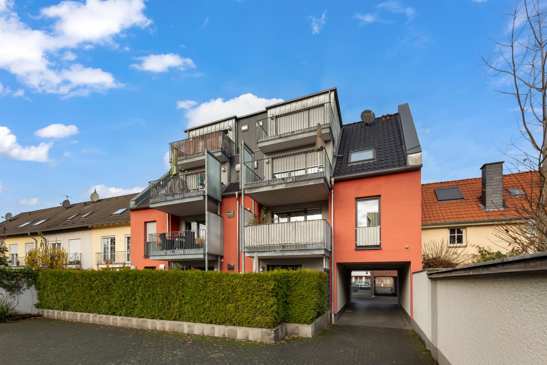 Modernes Mehrfamilienhaus mit Balkonen und Einfahrt, umgeben von Hecken und Nachbarhäusern.
