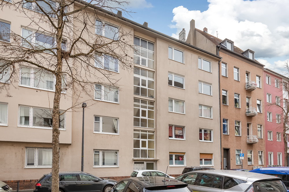 Mehrfamilienhaus mit mehreren Fenstern und geparkten Autos davor, kahle Bäume am Straßenrand.
