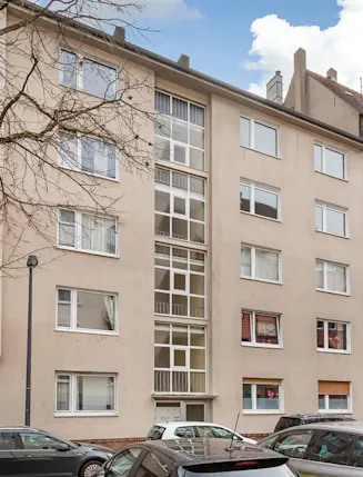 Mehrfamilienhaus mit mehreren Fenstern und geparkten Autos davor, kahle Bäume am Straßenrand.