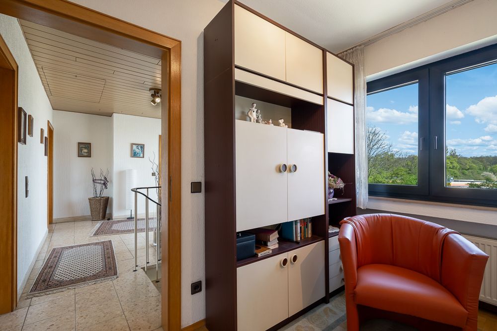 Helles Zimmer mit Regal, rotem Sessel und großem Fenster mit Blick ins Grüne.