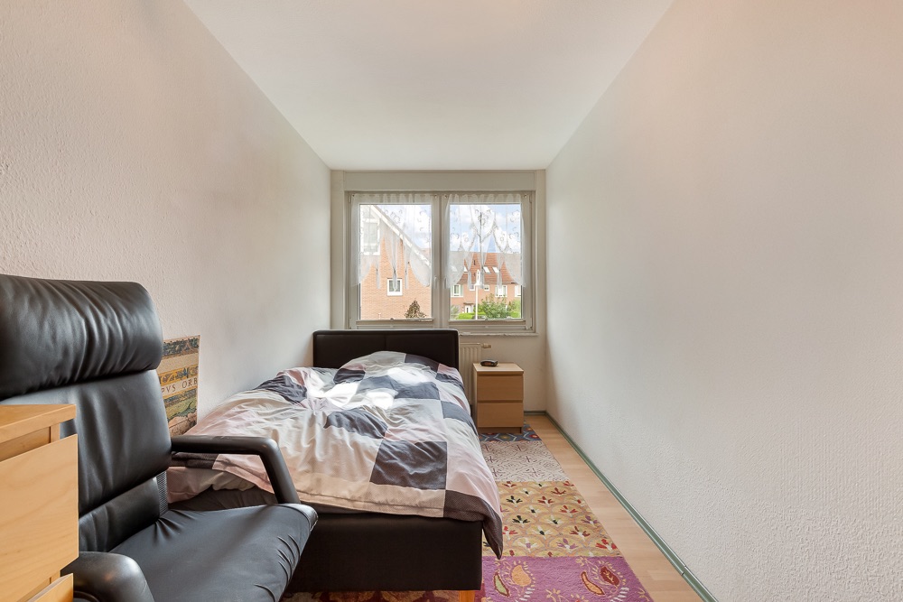 Schmales Schlafzimmer mit Einzelbett, Nachttisch und schwarzem Stuhl, Fenster am Raumende.