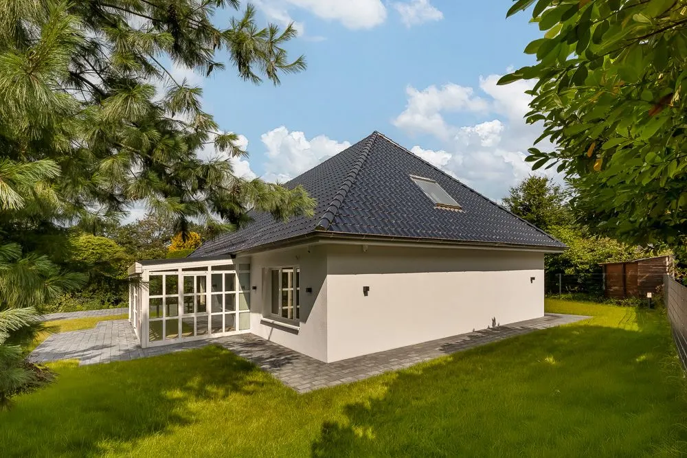 Einfamilienhaus mit dunklem Dach und Wintergarten, umgeben von grünem Garten und Bäumen.