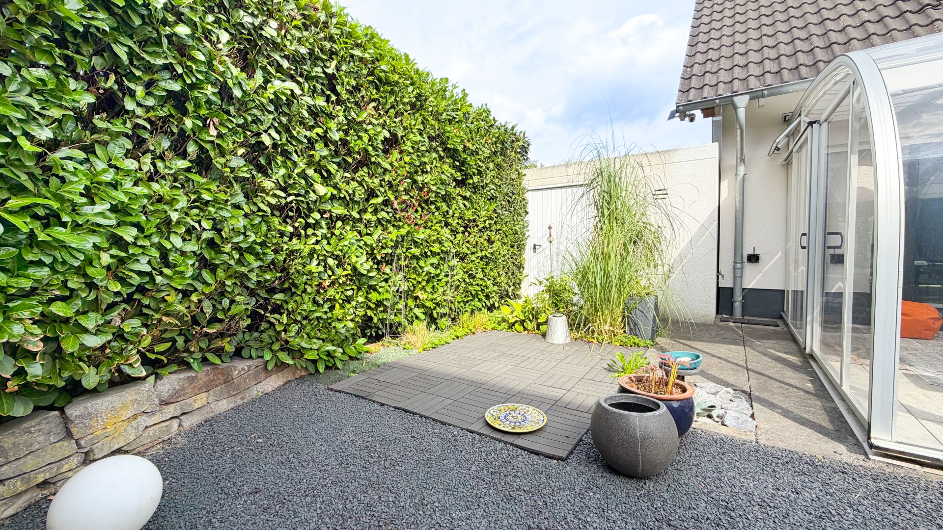 Kleiner Garten mit Kies, Grünpflanzen, Töpfen und Terrasse neben einem Wintergarten.