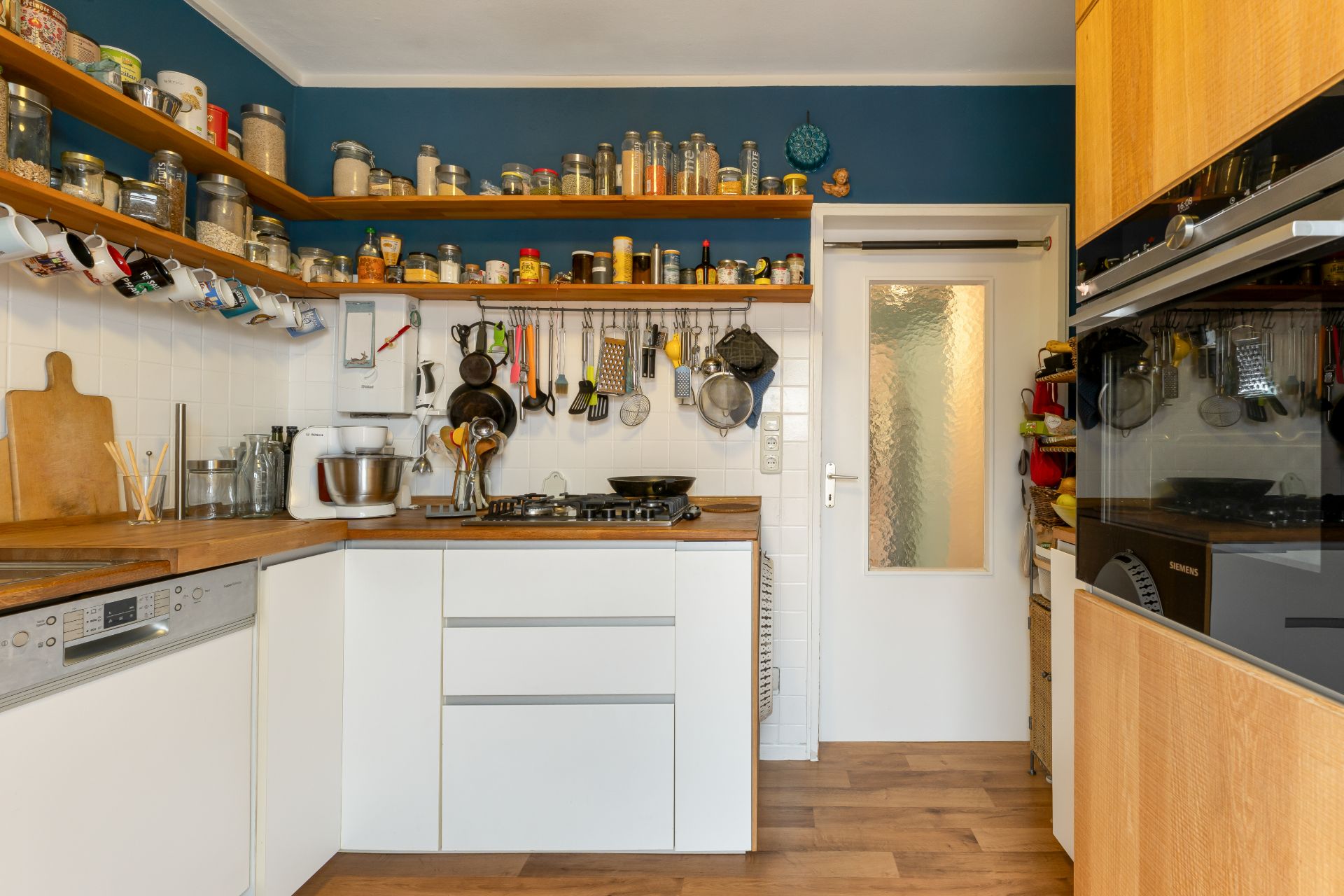 Moderne Wohnküche mit weißen Schränken, Holzarbeitsplatte und offenen Regalen voller Gläser und Küchenutensilien.