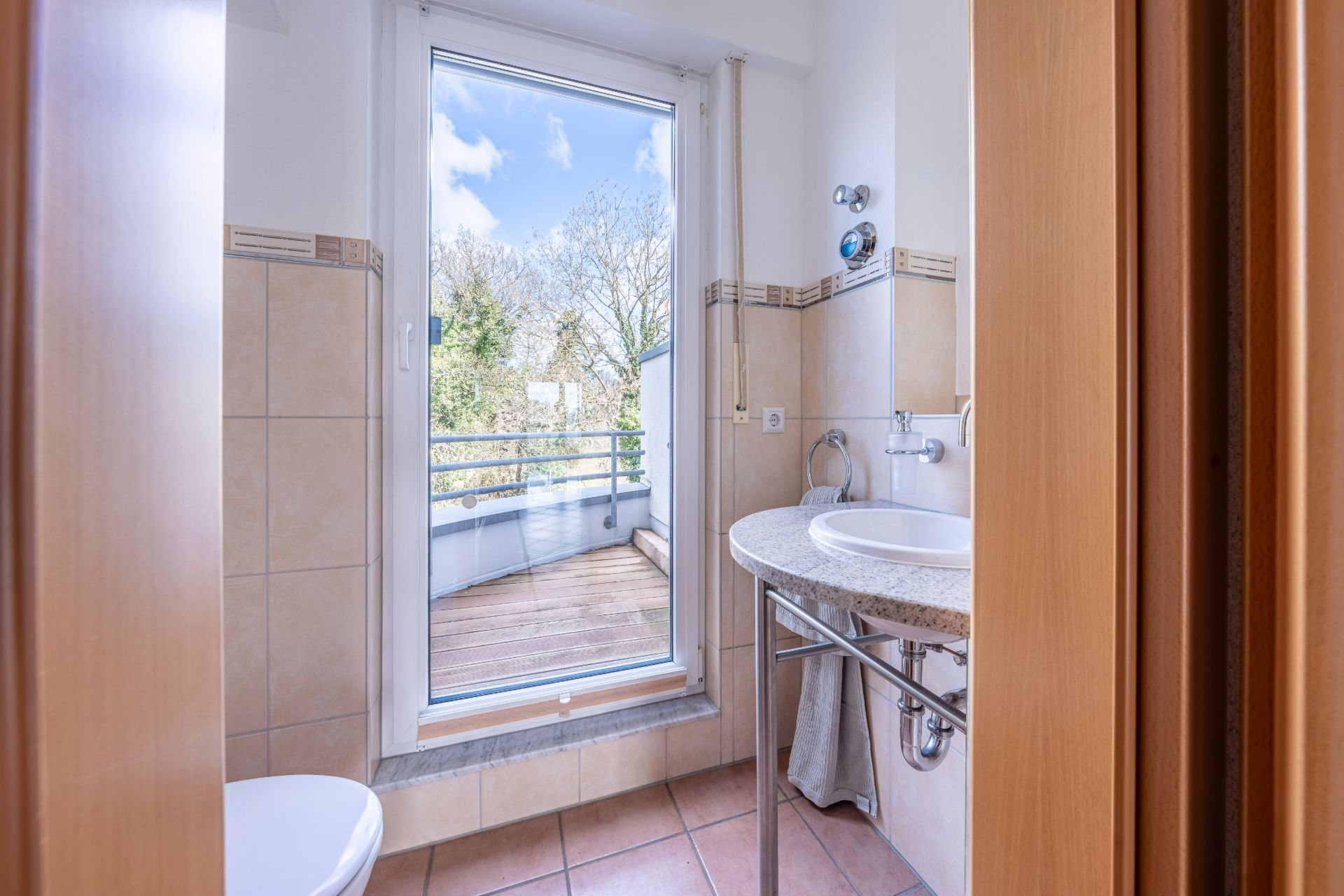 Helles Badezimmer mit Waschbecken, WC und Tür zu einem Balkon mit Holzfußboden.