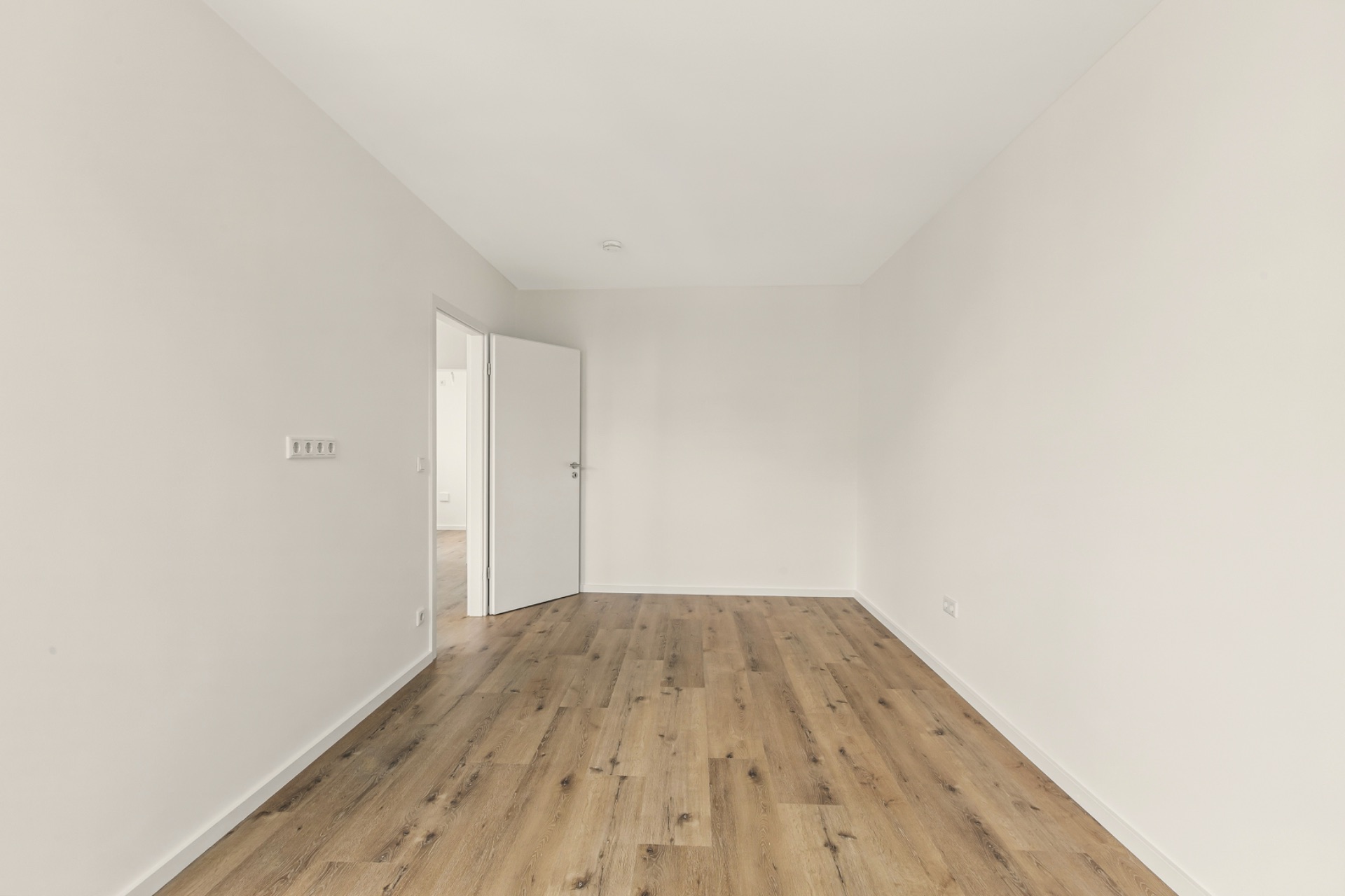 Leerer Raum mit hellen Wänden, Holzboden und offenstehender weißer Tür.