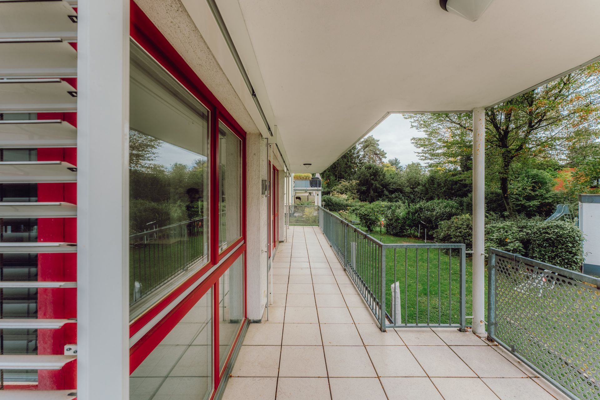 Überdachter Balkon mit Fliesenboden, Metallgeländer und Blick auf einen grünen Garten.