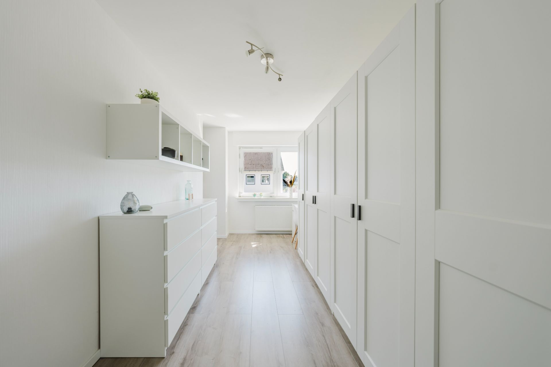 Heller Flur mit weißen Einbauschränken und Kommode, Fenster am Ende des Raumes.
