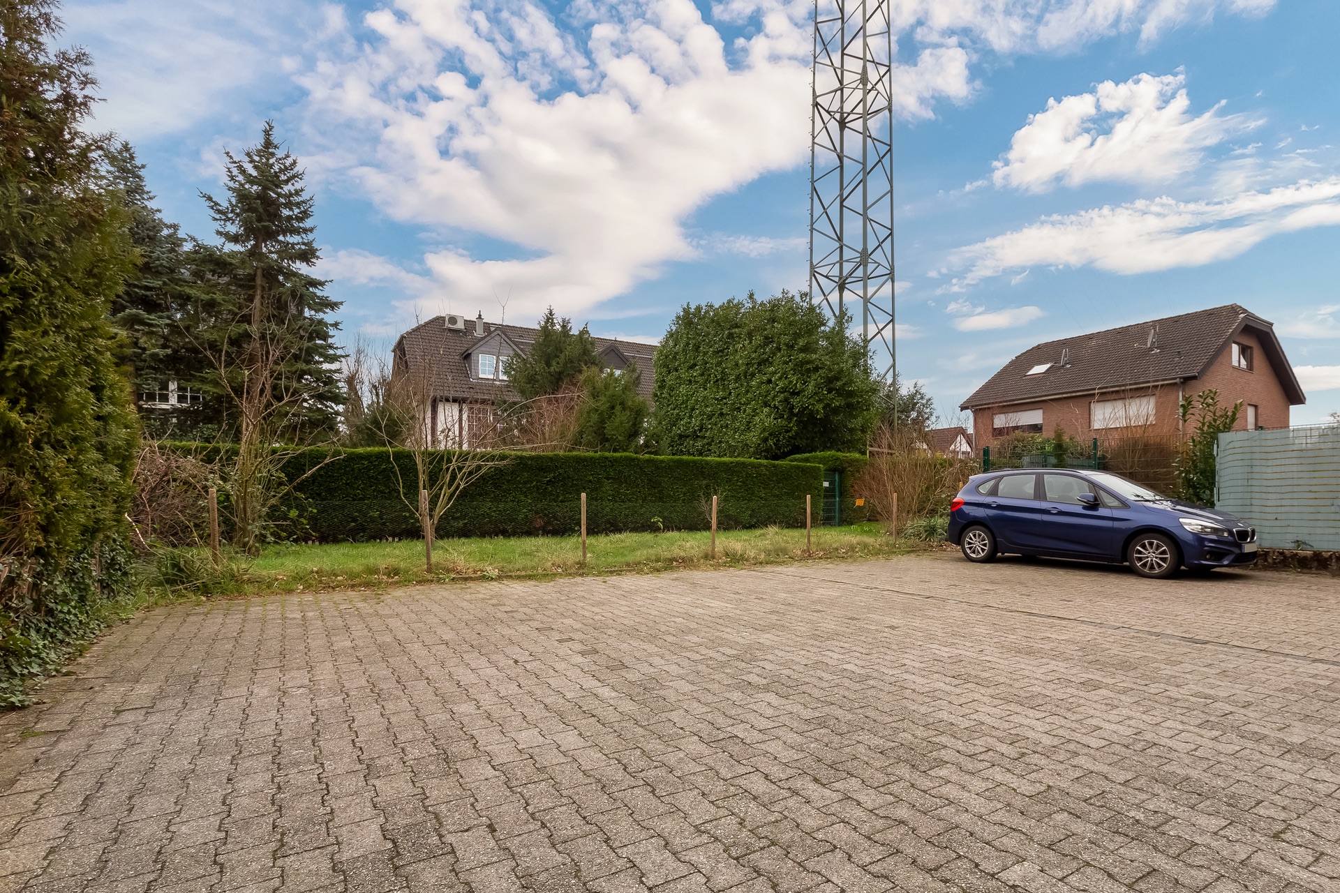 Gepflasterter Parkplatz mit einem blauen Auto, Wohnhäuser und Bäume im Hintergrund.