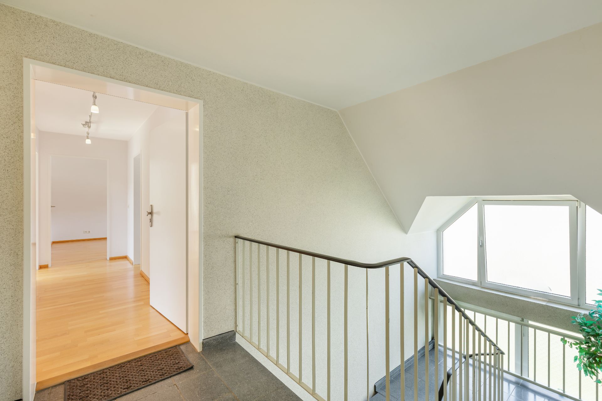Treppenhaus mit Geländer und Fenster, Tür führt in hellen Flur mit Parkettboden.