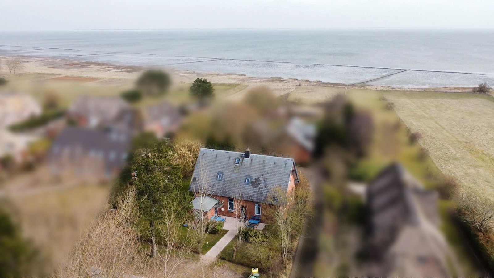 Backsteinhaus mit blauem Dach am Meer, von Bäumen und Feldern umgeben.