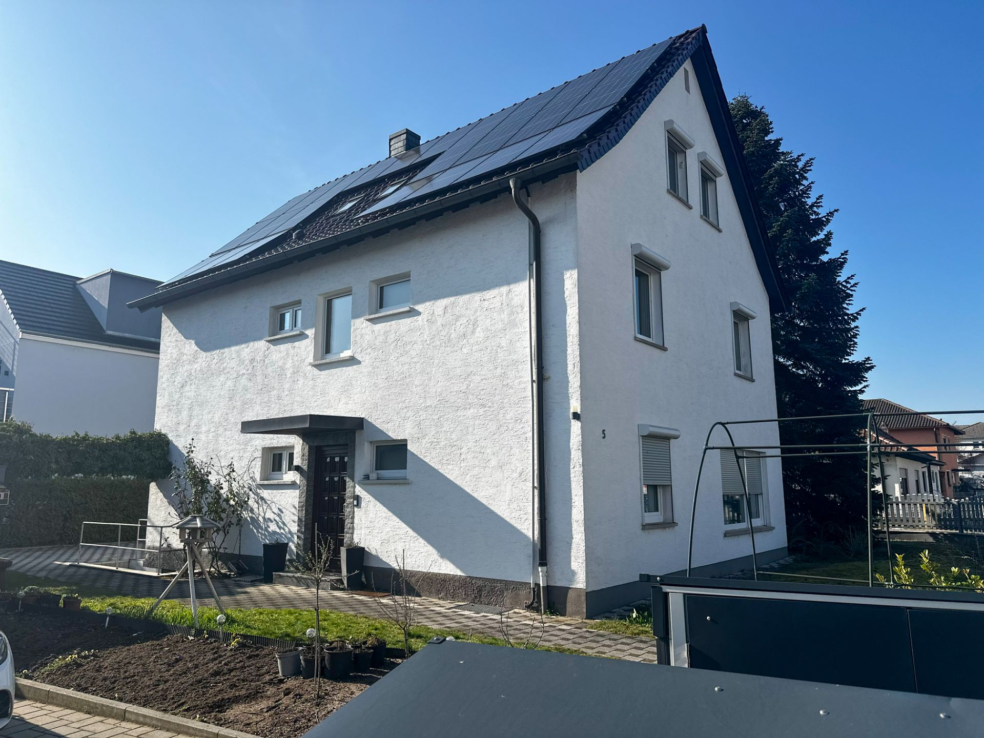 Weißes Einfamilienhaus mit dunklem Dach und Solarzellen, davor ein kleiner Garten.
