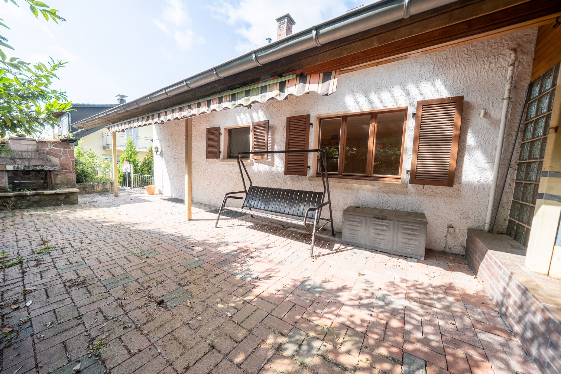 Gepflasterte Terrasse mit Hollywoodschaukel, Markise und Fensterläden an einem Wohnhaus.