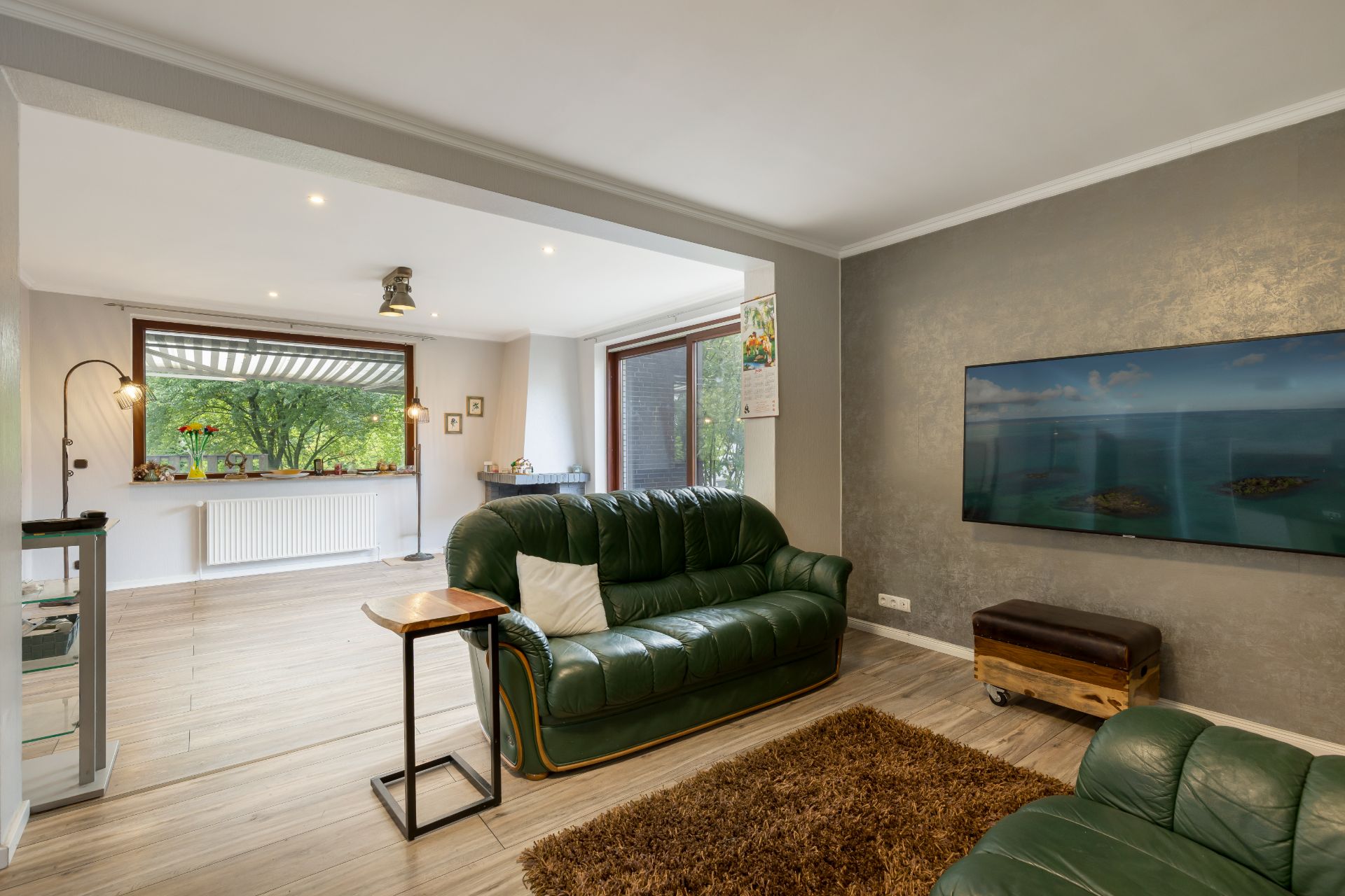 Helles Wohnzimmer mit grünen Ledersofas, Wandfernseher und großen Fenstern mit Blick ins Grüne.