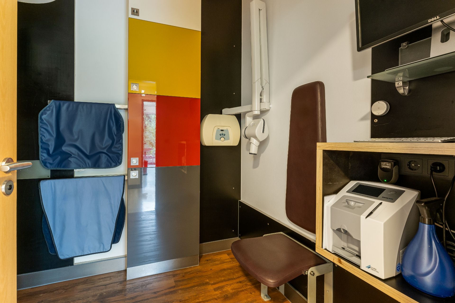 Kleiner Raum mit braunem Sitz, farbiger Tür, Computer und medizinischen Geräten.