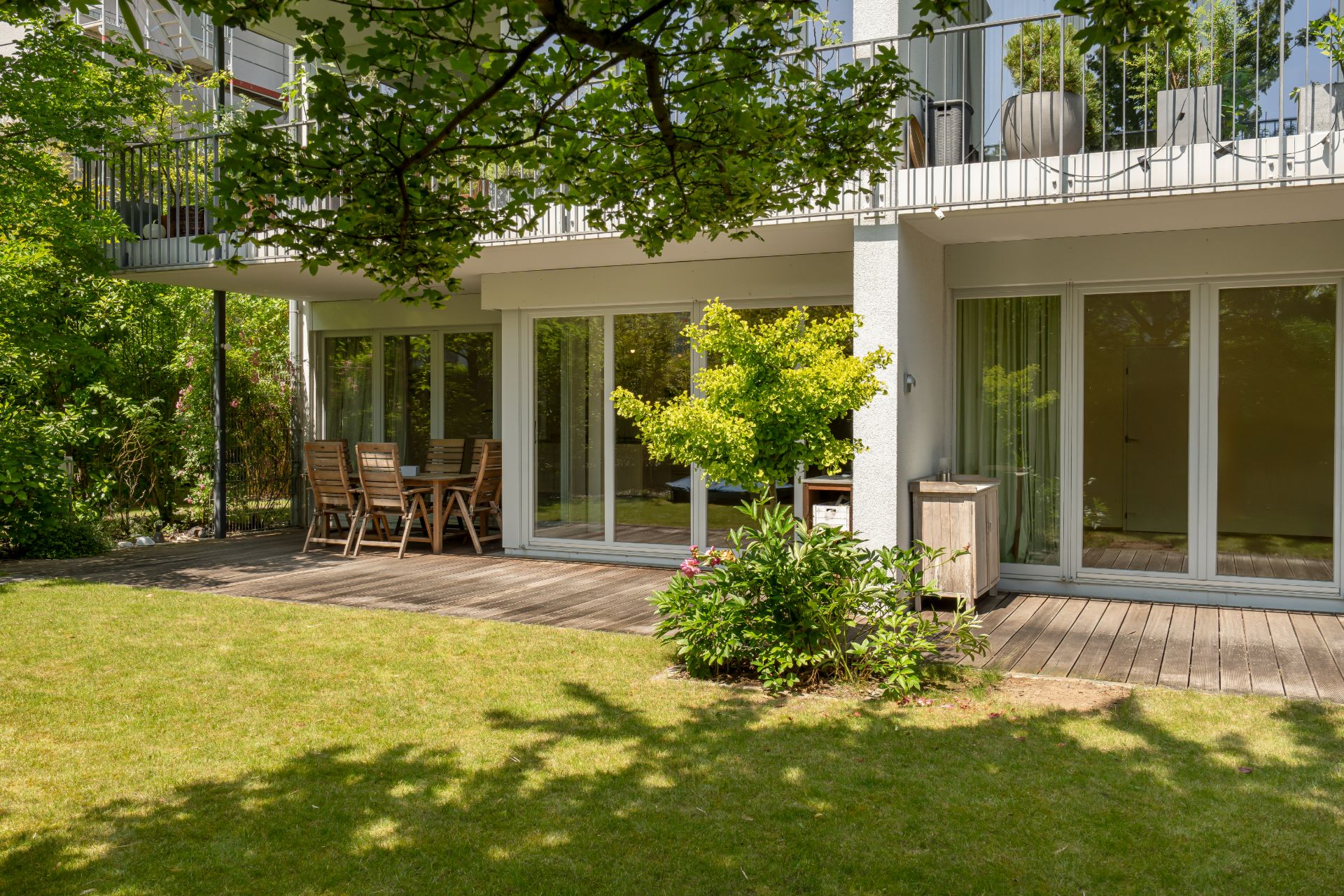 Terrasse mit Holzboden, Gartentisch und Stühlen vor einer modernen Wohnung mit großen Glasfenstern.