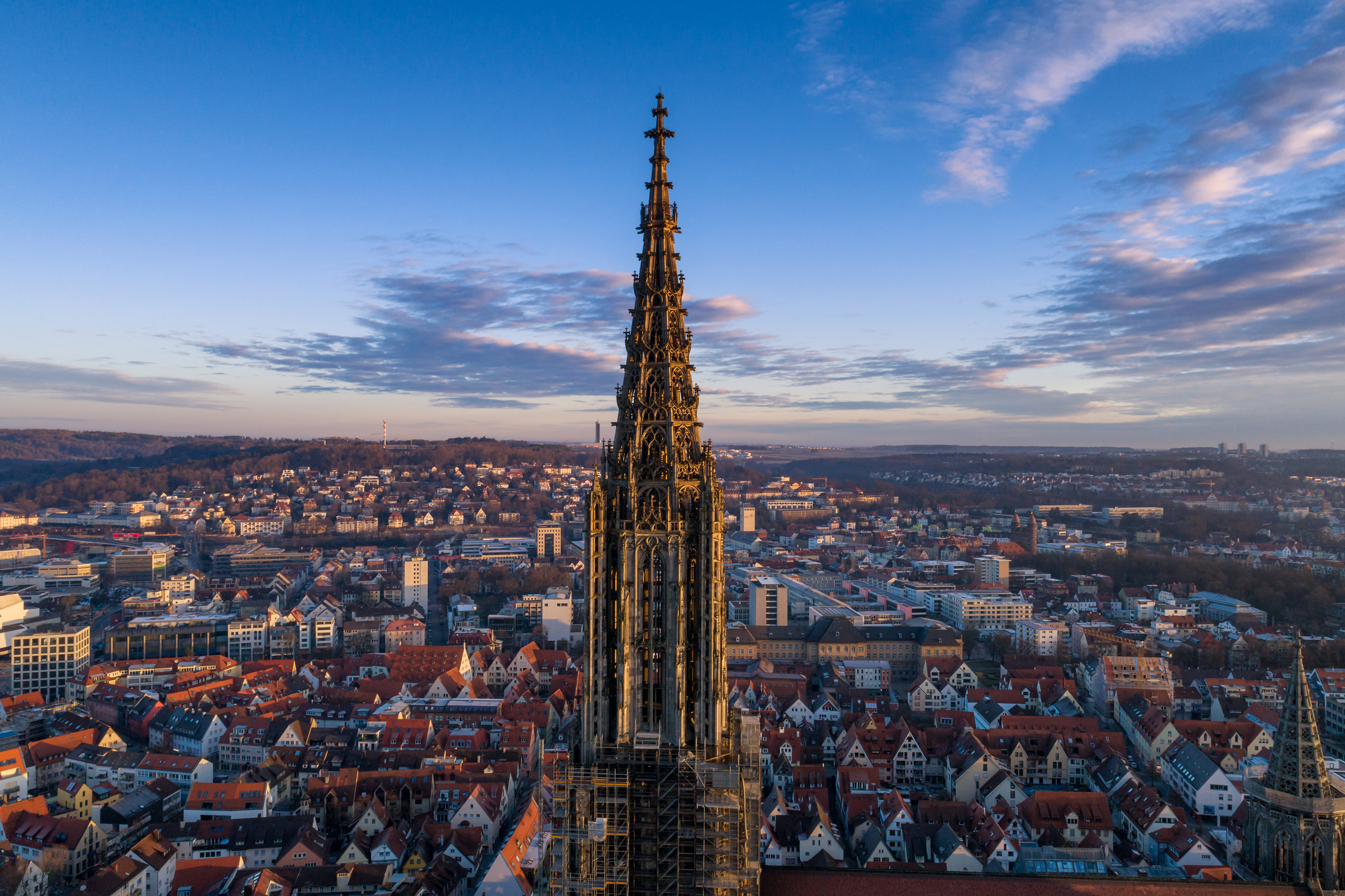 Spitze eines hohen Kirchturms mit Stadt im Hintergrund bei blauem Himmel.