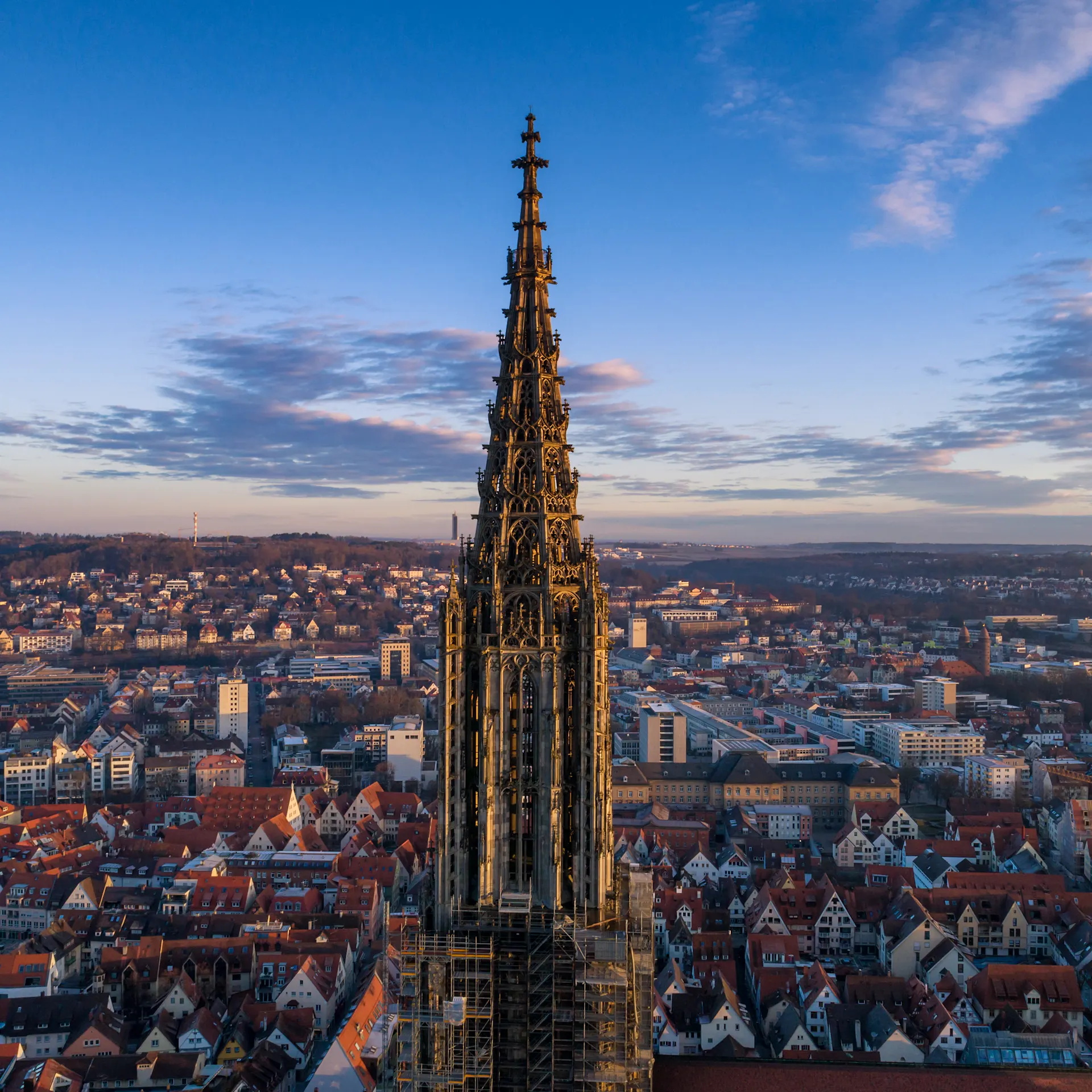 Spitze eines hohen Kirchturms mit Stadt im Hintergrund bei blauem Himmel.