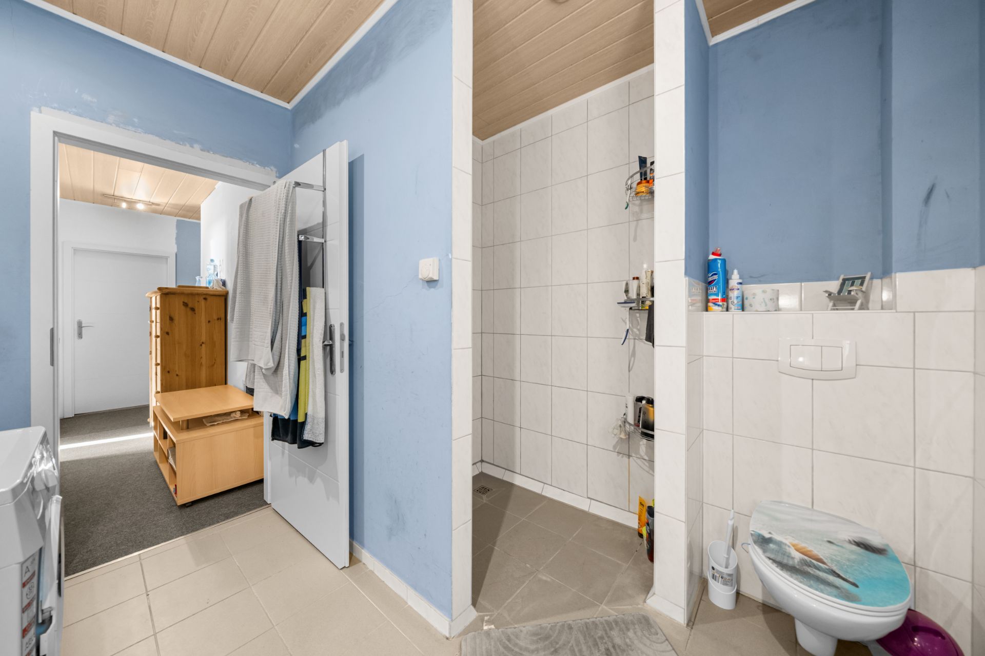 Badezimmer mit offener Dusche, Toilette und hellblauen Wänden, daneben Flur mit Holzbank.
