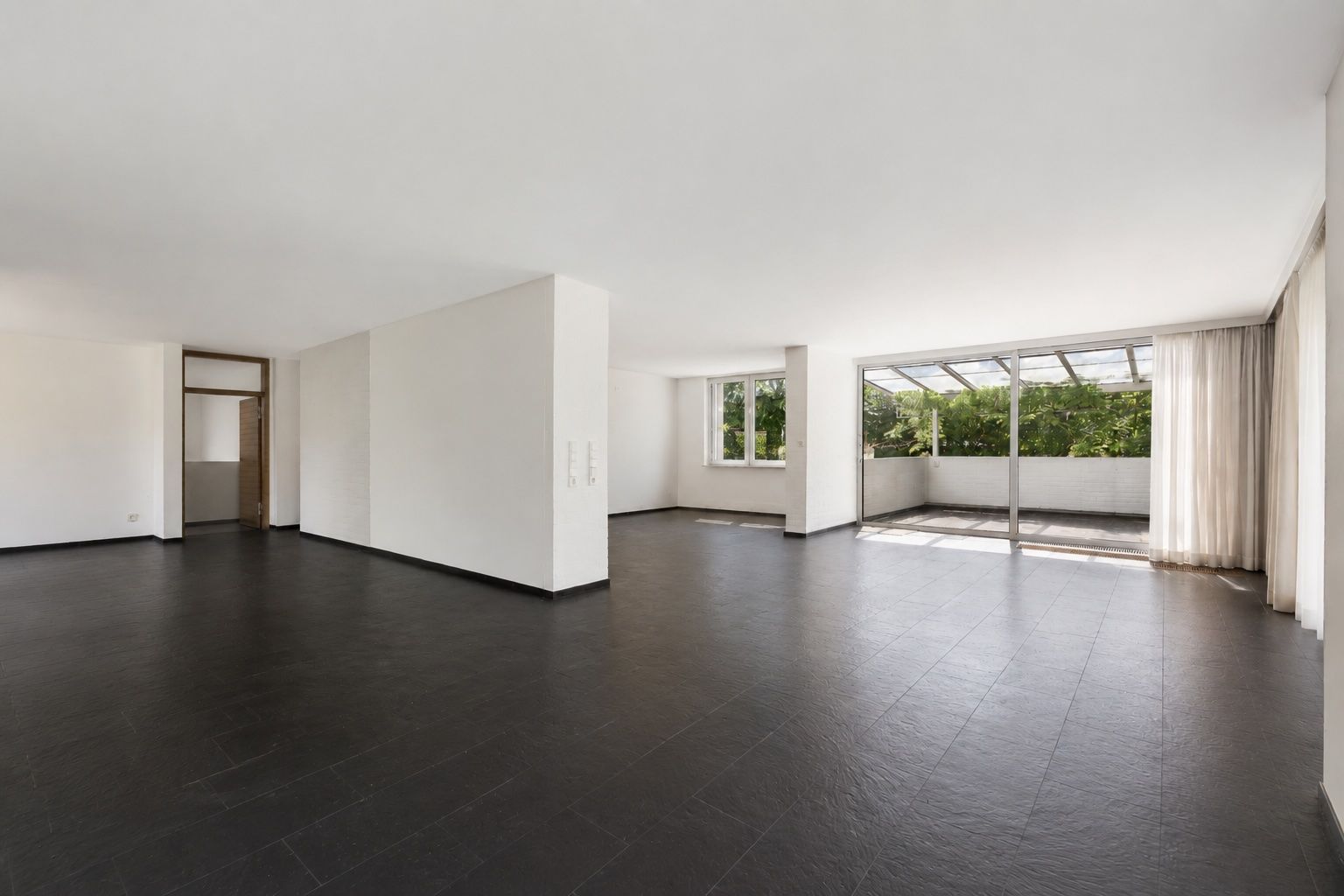Großer, leerer Wohnraum mit dunklem Boden, weißen Wänden und Zugang zu einer Terrasse mit Glasdach.