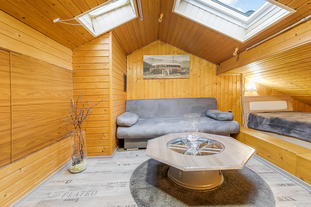 Gemütlicher Dachraum mit Holzverkleidung, grauem Sofa, Bett und Couchtisch unter zwei Dachfenstern.