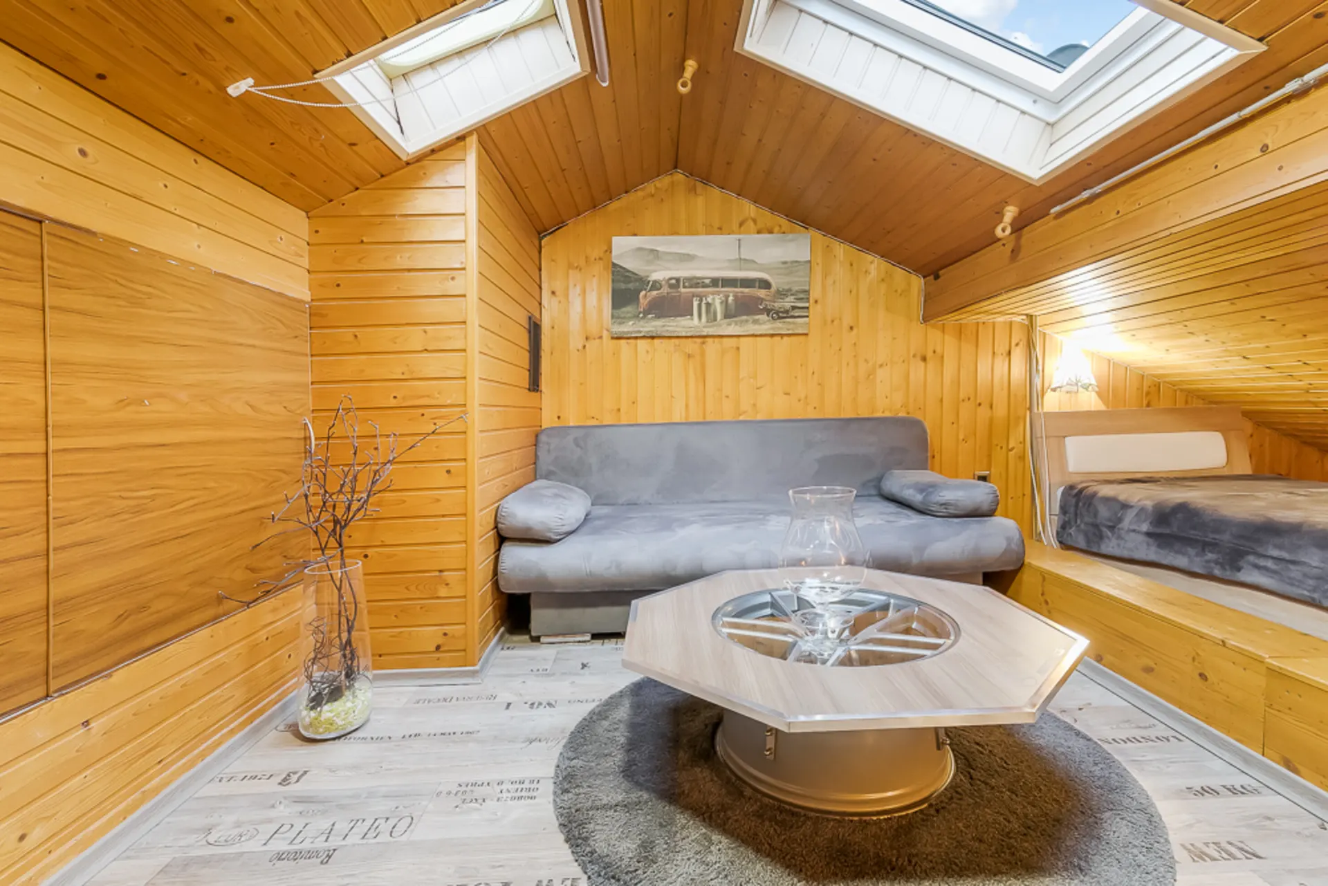 Gemütlicher Dachraum mit Holzverkleidung, grauem Sofa, Bett und Couchtisch unter zwei Dachfenstern.