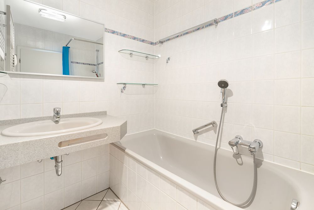 Helles Badezimmer mit Badewanne, Waschbecken, Spiegel und weißen Fliesen.