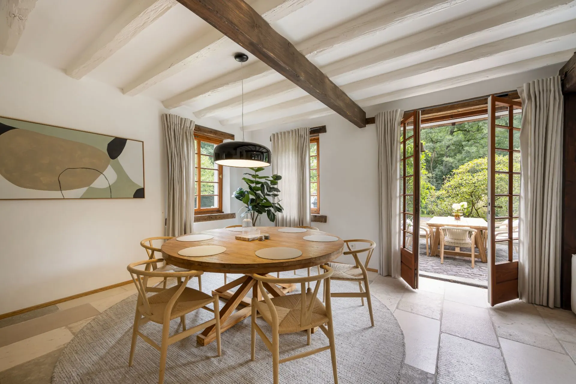 Esszimmer mit rundem Holztisch, sechs Stühlen, großen Fenstern und geöffneten Türen zur Terrasse.