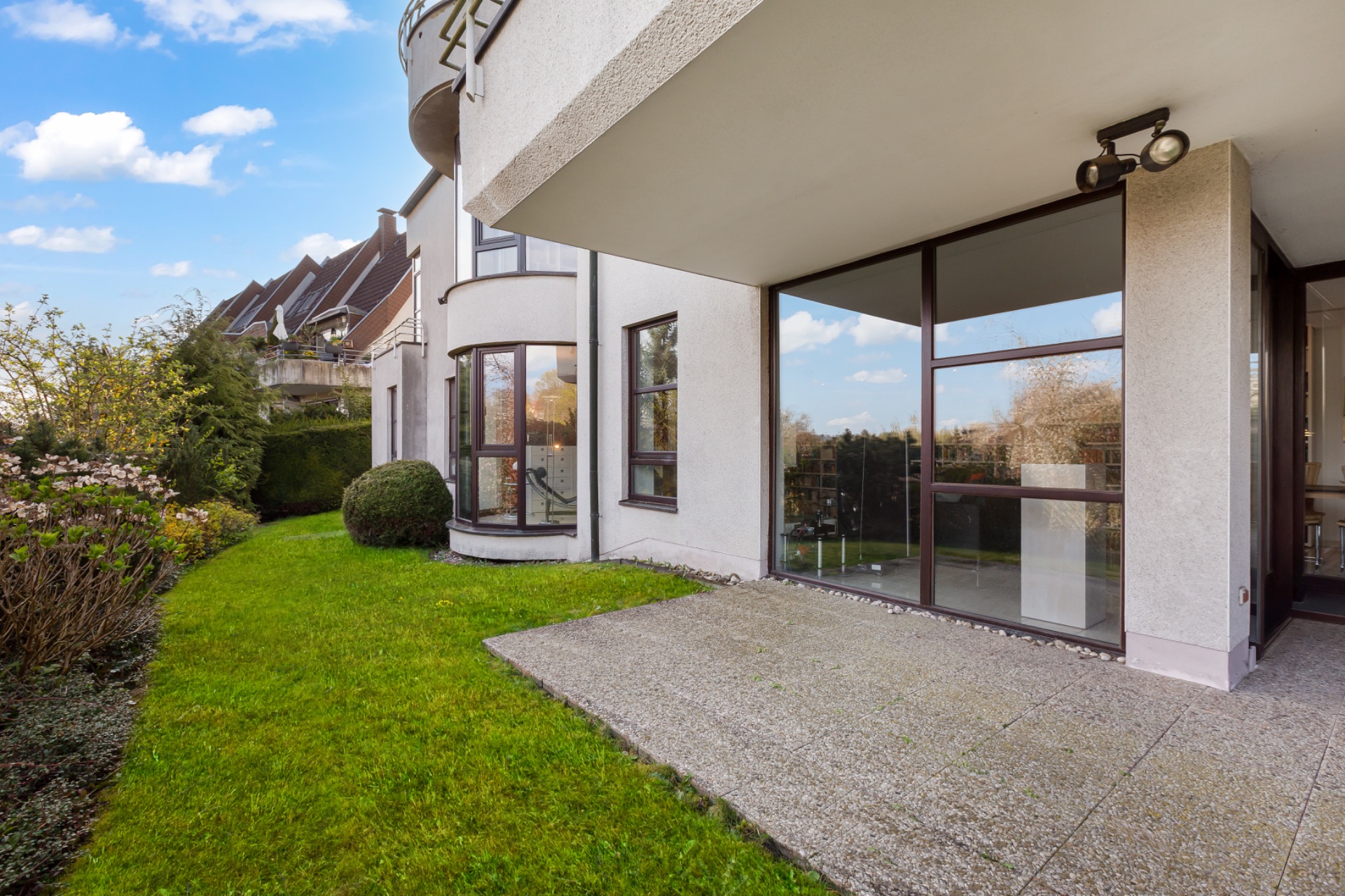 Moderne Terrasse mit Glasfassade und Garten, daneben grüne Hecke und Blumenbeet.