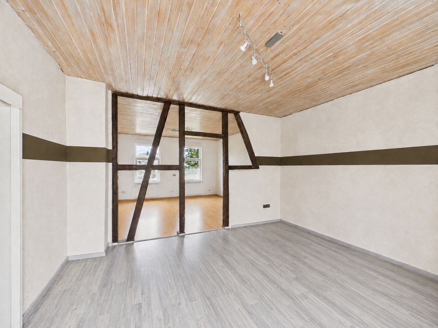 Leerer Raum mit Holzbalken an der Wand und Blick in ein weiteres Zimmer mit Fenster.