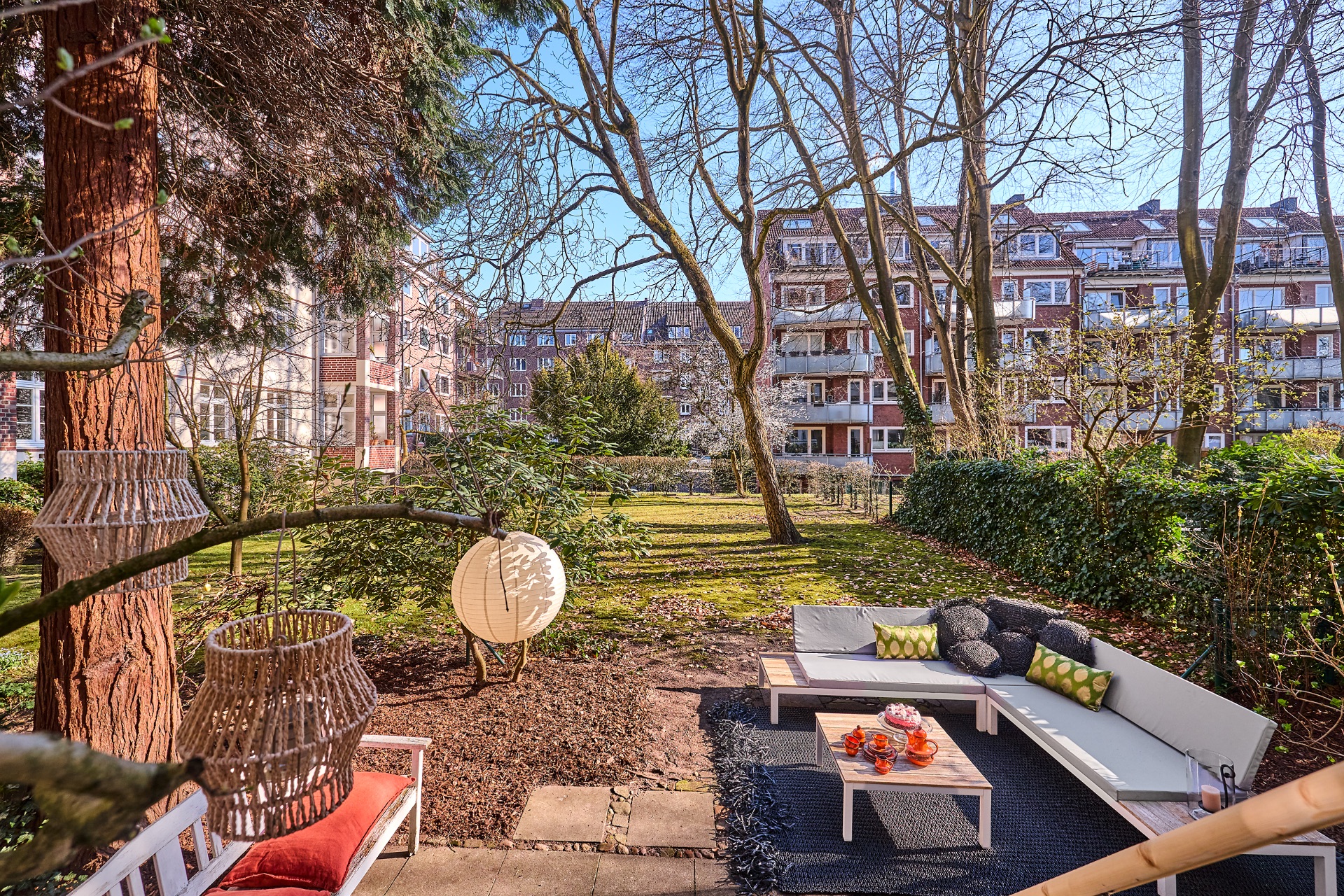 Garten mit Sitzbereich, Sofas, Couchtisch und hängenden Laternen, umgeben von Bäumen und Wohnhäusern.