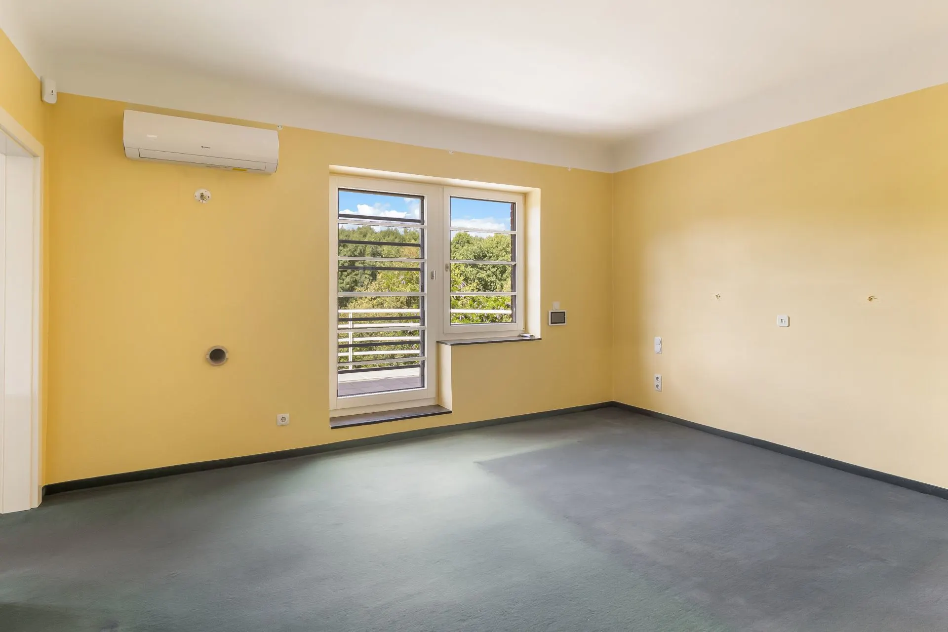 Leeres Zimmer mit gelben Wänden, großem Fenster und Klimaanlage an der Wand.