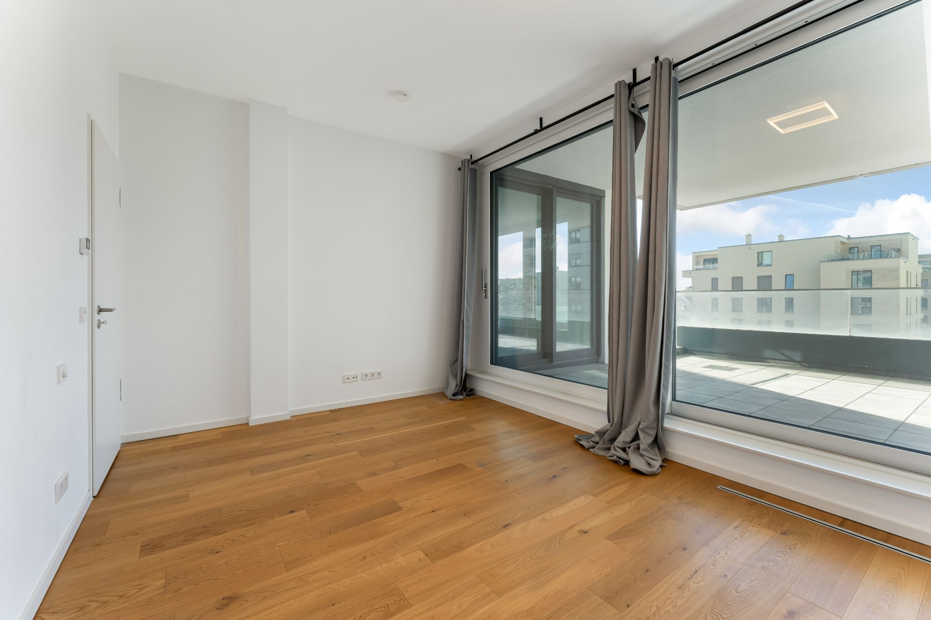 Leeres Zimmer mit Holzboden, großen Fenstern und Zugang zu einem Balkon.