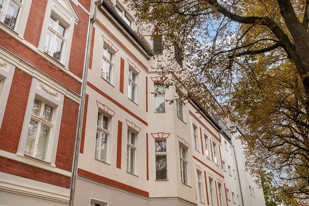 Mehrstöckiges Wohngebäude mit roten und weißen Fassadenelementen, daneben Bäume mit Herbstlaub.