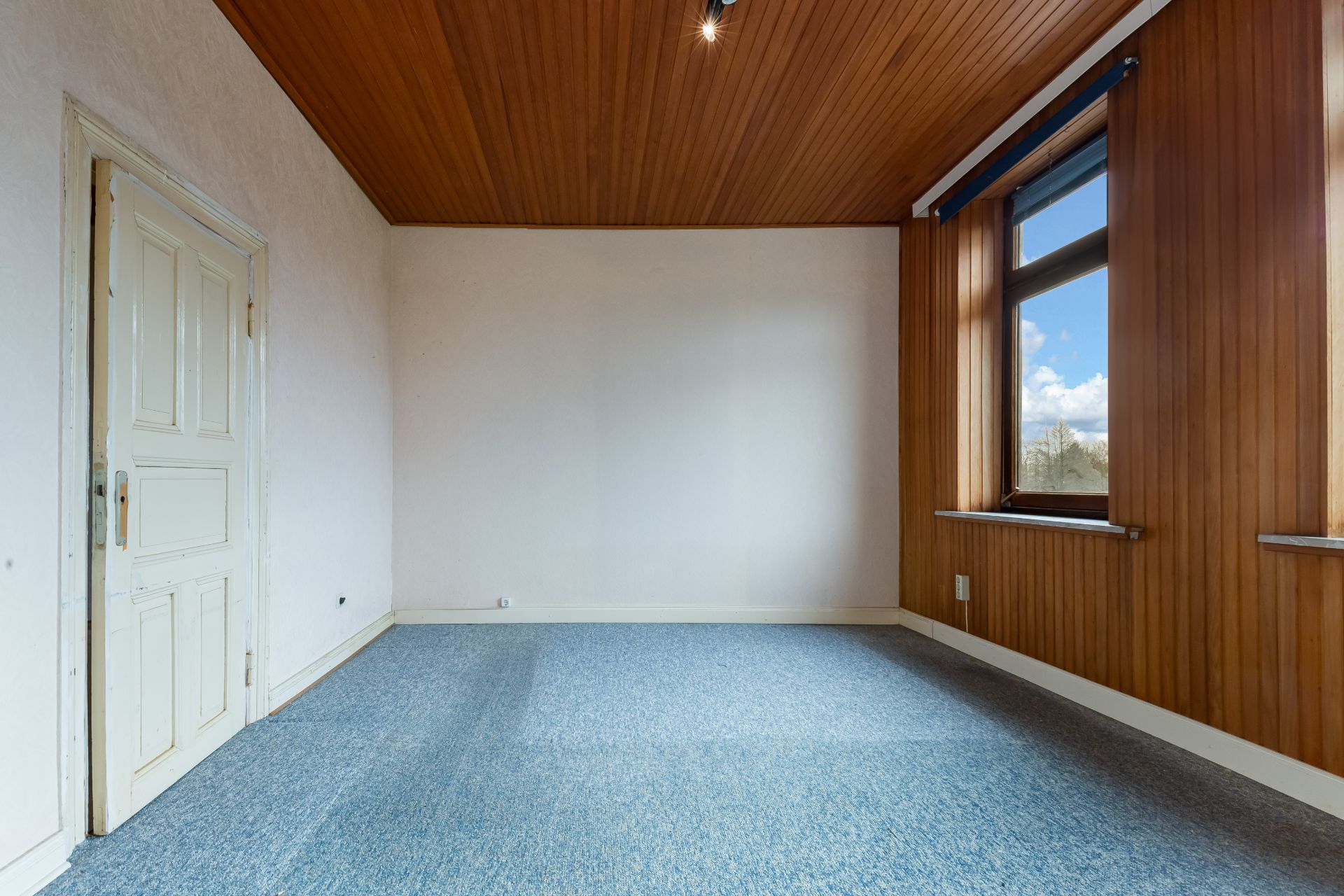 Leeres Zimmer mit blauer Auslegware, Holzdecke, holzvertäfelten Wänden und großem Fenster.