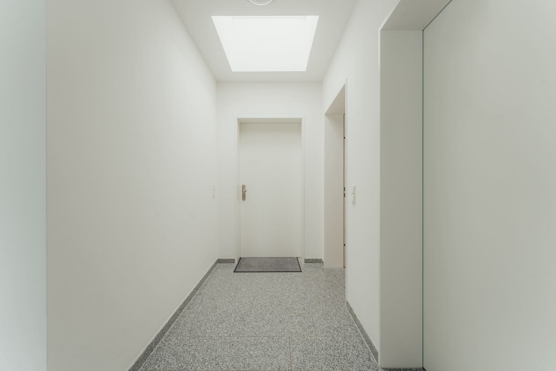 Heller Flur mit grauem Boden, weißer Tür und rechteckigem Dachfenster.