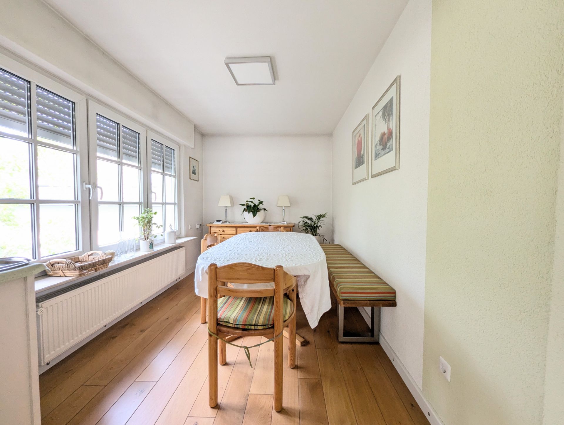 Esszimmer mit großem Fenster, Tisch, Stühlen, Bank, Pflanzen und Bildern an der Wand.