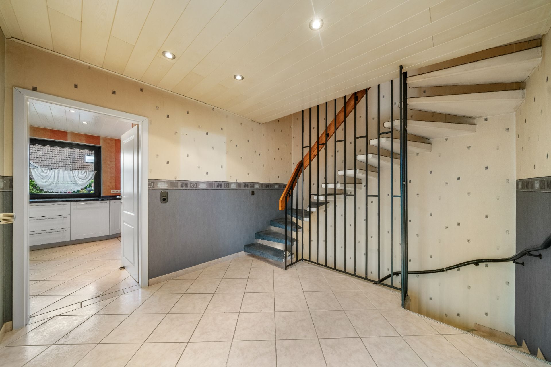 Flur mit gefliester Treppe, hellem Boden und Zugang zu einer Küche mit Fenster.
