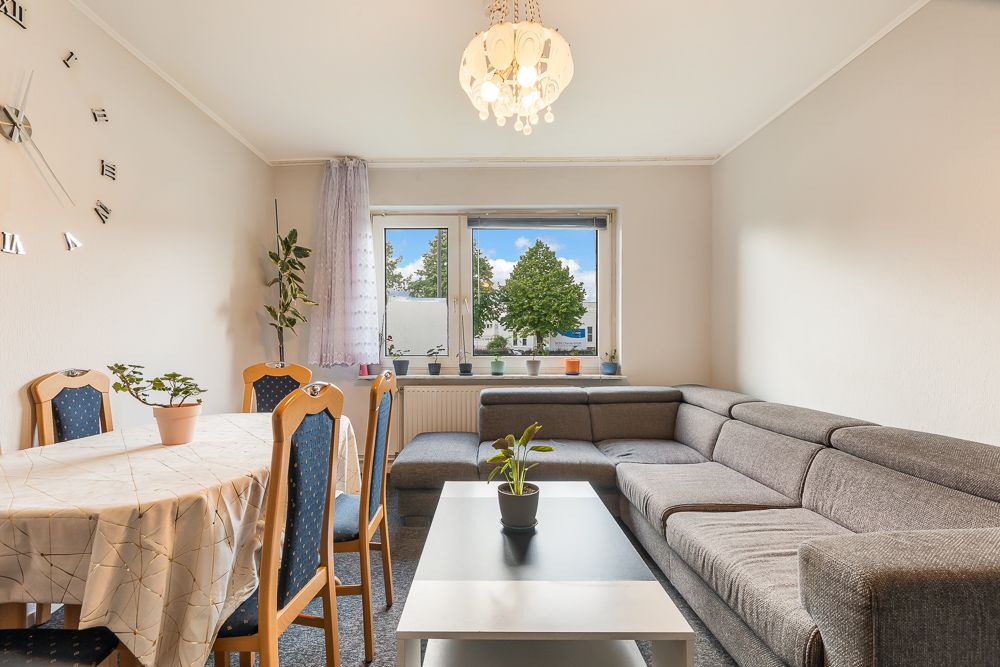 Wohnzimmer mit grauer Eckcouch, Esstisch, Holztischstühlen, Zimmerpflanzen und großem Fenster.