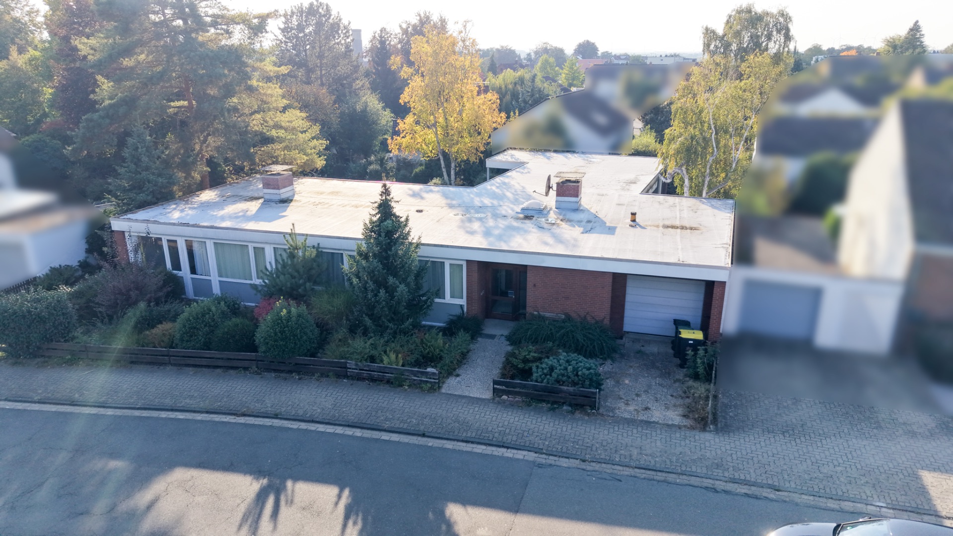 Einfamilienhaus mit Flachdach, großem Fenster und Garage, umgeben von Bäumen und Sträuchern.