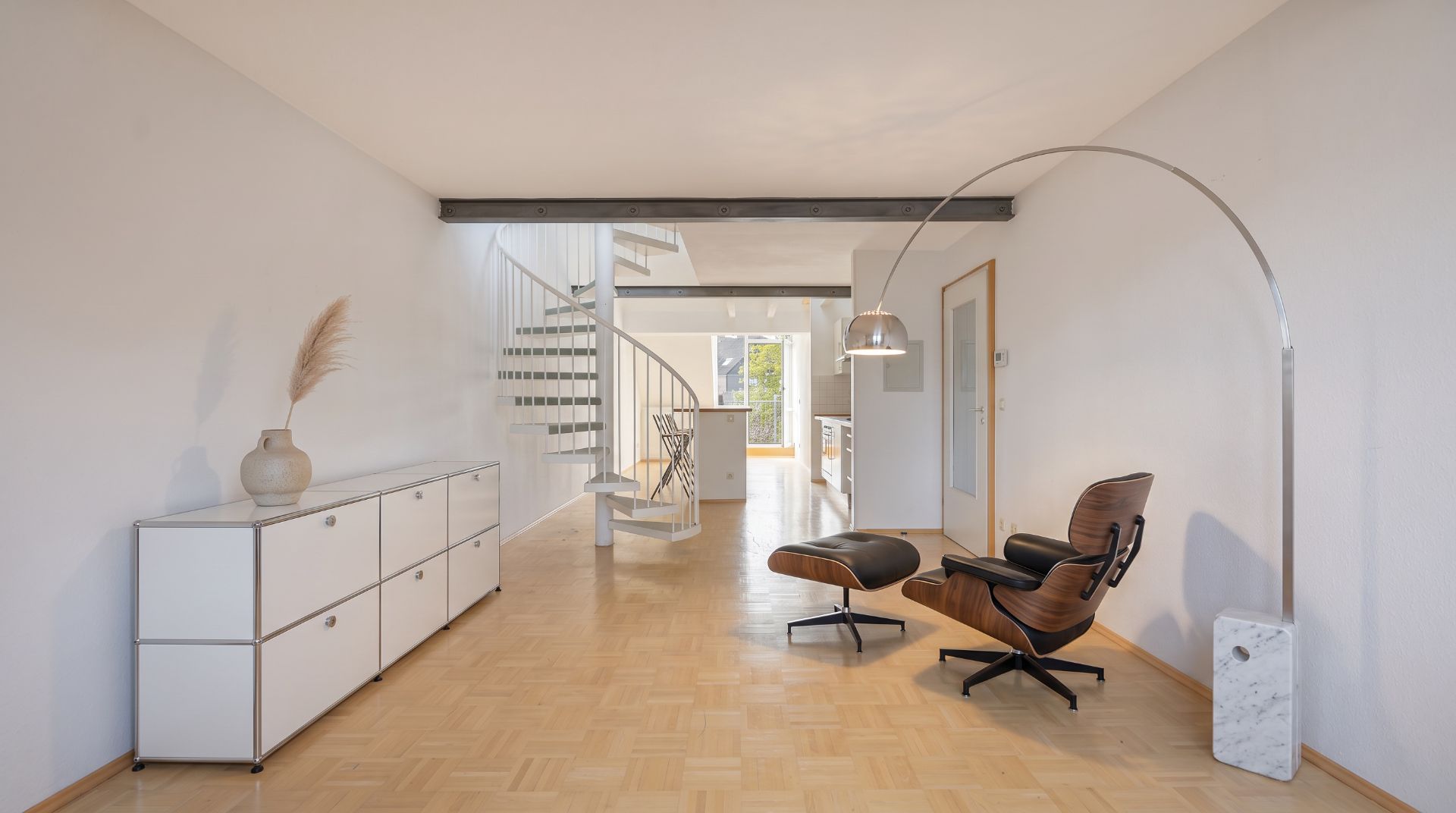 Heller Wohnraum mit Holzfußboden, Sessel, Stehlampe, Kommode und einer weißen Wendeltreppe.