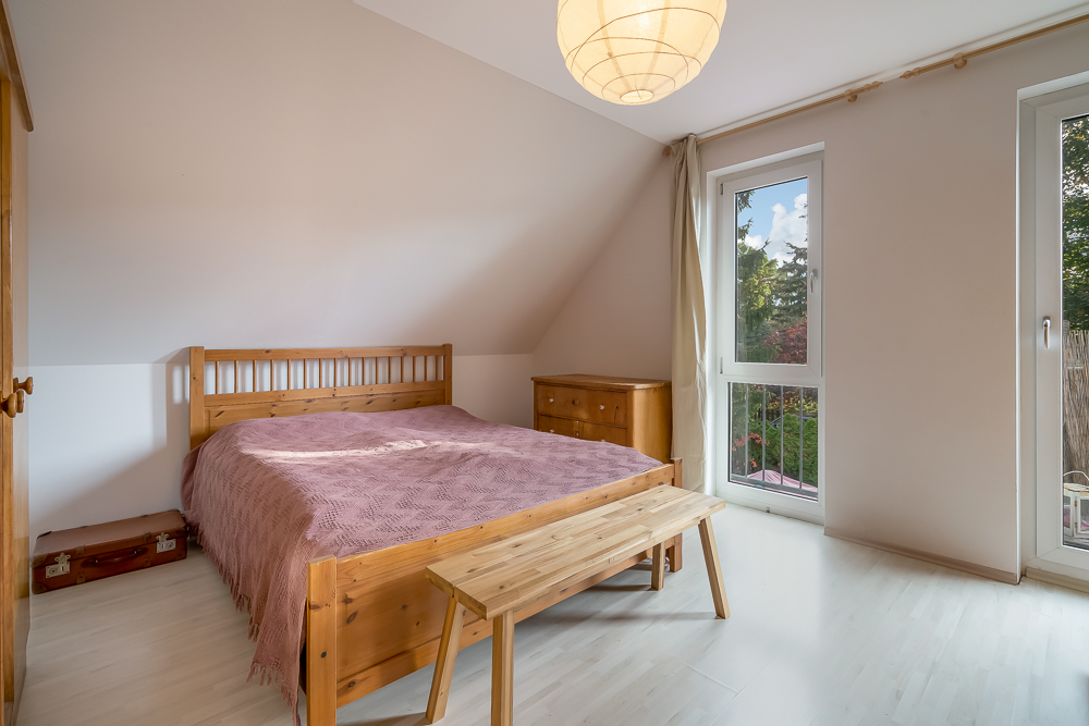 Helles Schlafzimmer mit Holzbett, Bank, Kommode und zwei großen Fenstern.