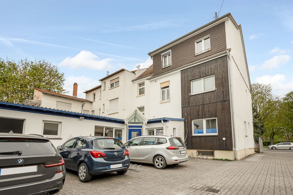 Mehrstöckiges Wohnhaus mit mehreren geparkten Autos auf einem gepflasterten Parkplatz davor.