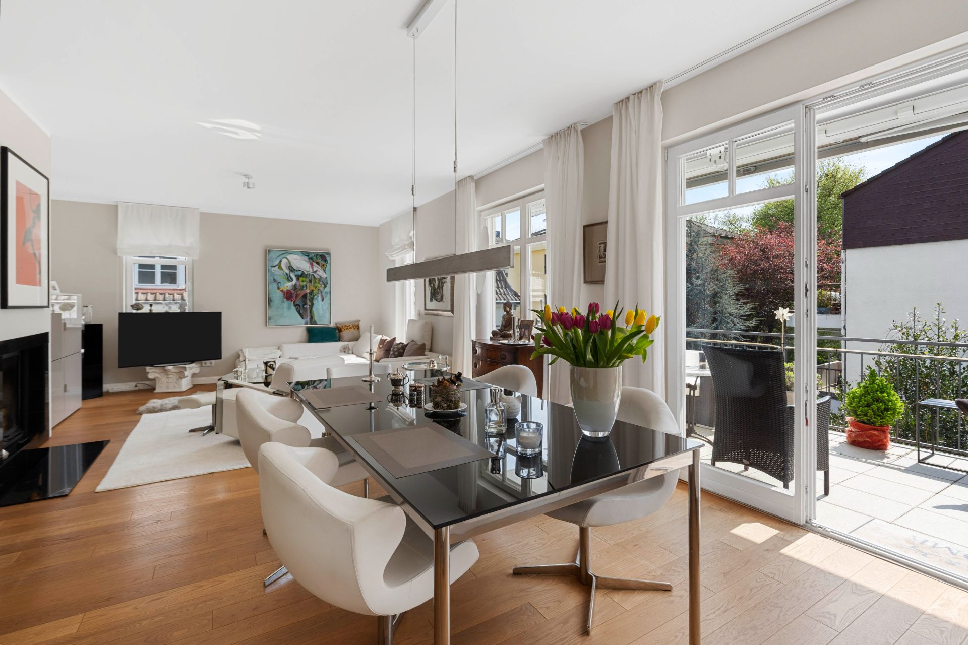Helles Wohnzimmer mit Esstisch, bequemen Stühlen, Blumen auf dem Tisch und Zugang zum Balkon.