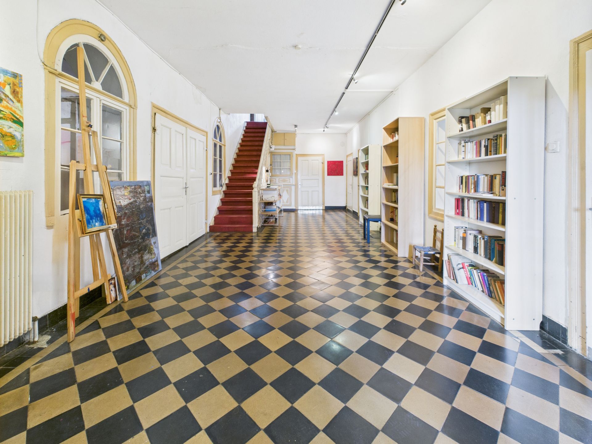 Großer Flur mit schwarz-beigem Schachbrettboden, roten Treppen, Bücherregalen und Staffelei.