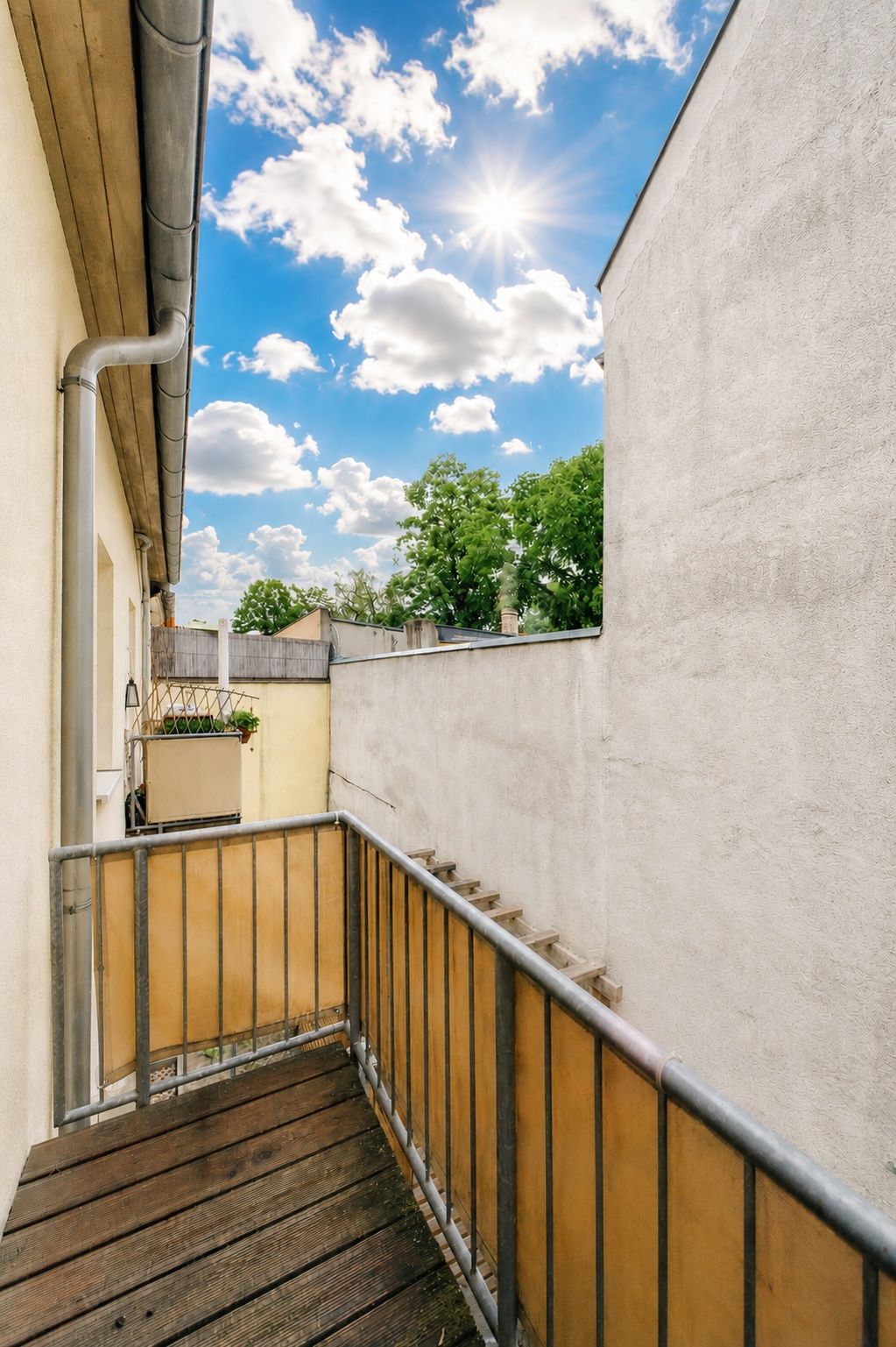 Kleiner Balkon mit Holzboden und Metallgeländer, Blick auf Himmel und Bäume.