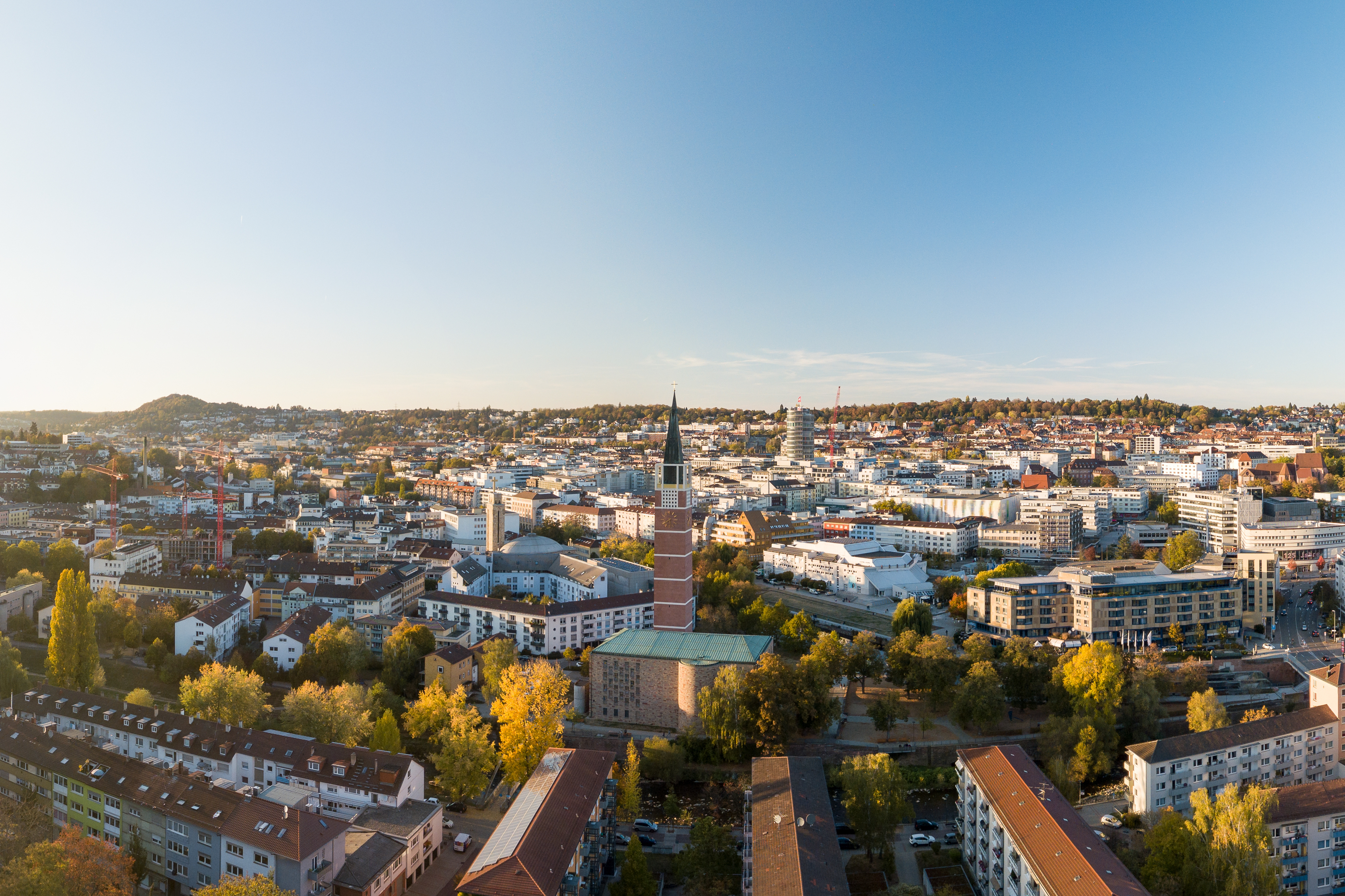 Stadtpanorama mit Wohnhäusern, Bäumen und einer Kirche mit hohem Turm im Vordergrund.