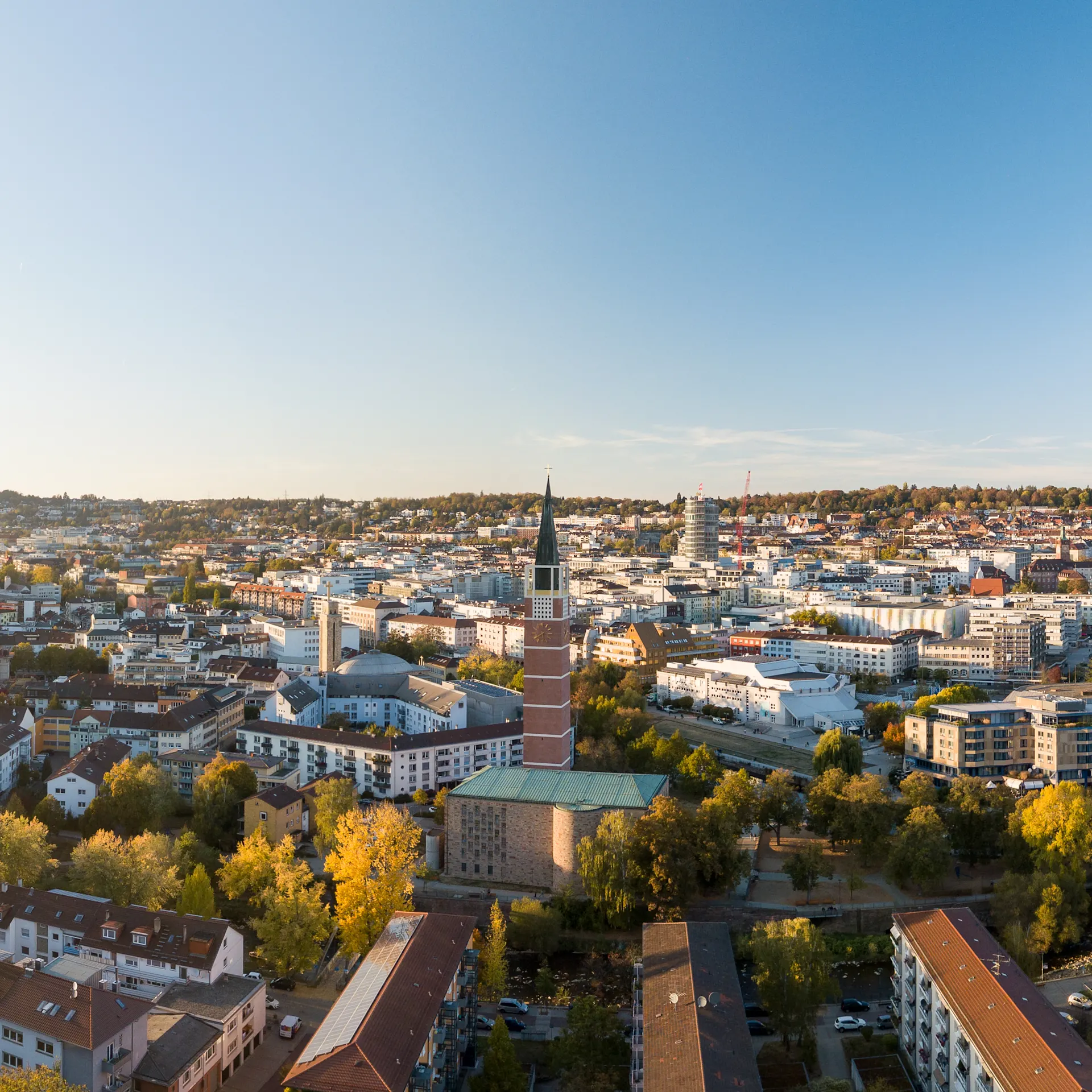 Stadtpanorama mit Wohnhäusern, Bäumen und einer Kirche mit hohem Turm im Vordergrund.