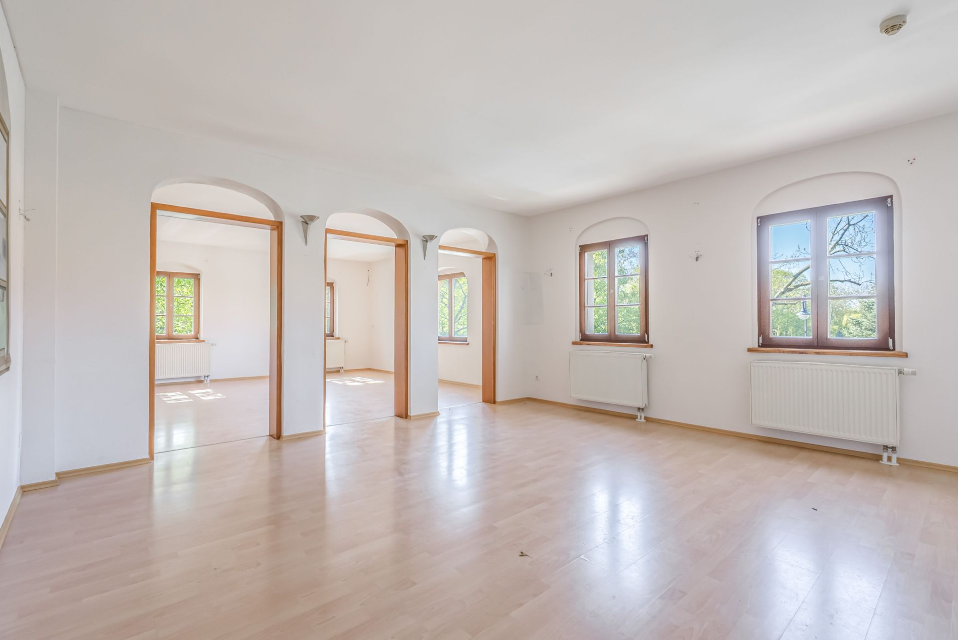 Helles, leeres Zimmer mit großen Fenstern, drei Rundbögen und Parkettboden.
