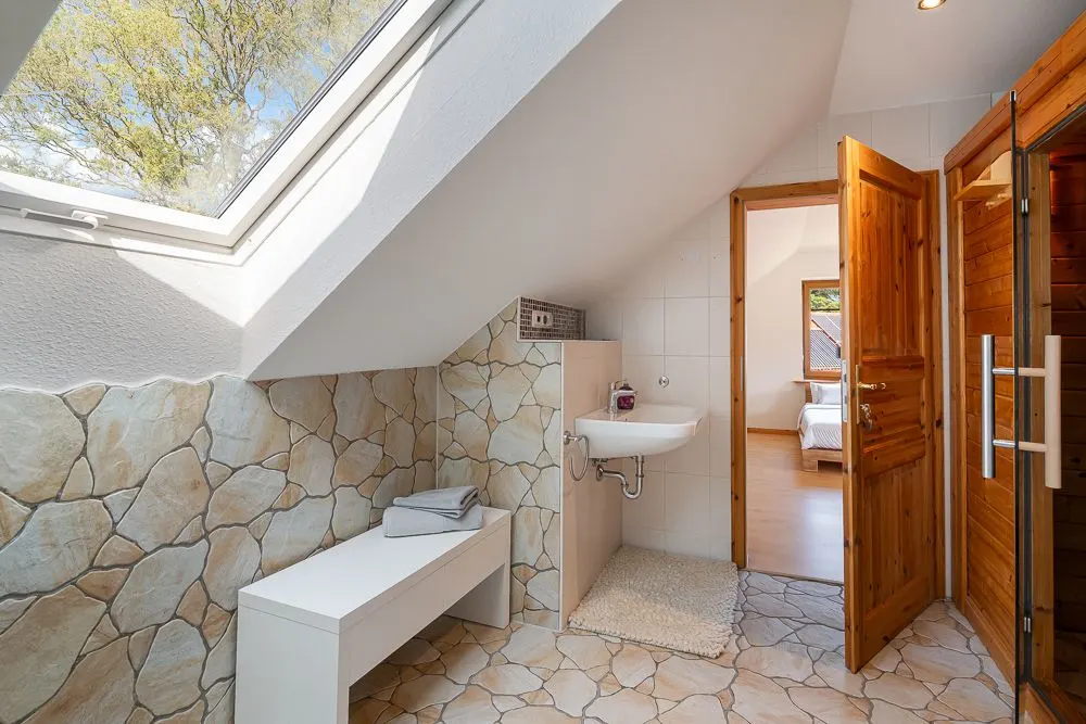 Helles Bad mit Dachfenster, Steinfliesen, Waschbecken, Bank und Zugang zum Schlafzimmer.